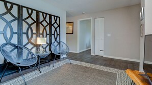 A modern living room with two black wireframe chairs, a small table with a lamp, a decorative wall panel, and a wooden bench. - 15 Mins to Closest Beach, 10 minutes from Downtown, with FIrepit! (Tampa)