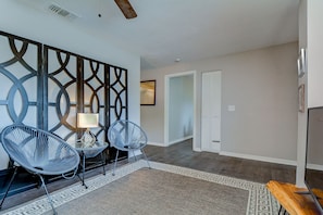 A modern living room with two black wireframe chairs, a small table with a lamp, a decorative wall panel, and a wooden bench.