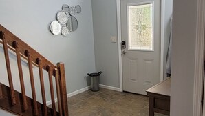 A spacious hallway with a wooden staircase, a chandelier, a door with a glass panel, and a trash can.