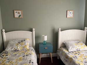 A bedroom with two white beds, floral bedding, a small blue nightstand with a lamp, and two framed pictures on the wall.