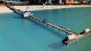 An aerial view of a coastal area with a pier extending into turquoise waters, a sandy beach, and a row of buildings in the background. - Beautiful home in Oldsmar (Oldsmar)
