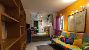 A cozy living room with a colorful sofa, wooden shelves, a kitchen area in the background, and a large framed map on the wall.