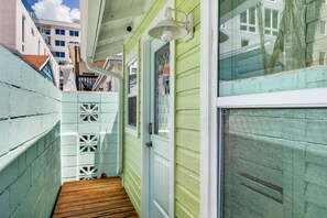 Une étroite terrasse en bois mène à une porte dotée d’une fenêtre, le tout étant entouré de murs vert clair et d’une clôture blanche ornée de motifs décoratifs.