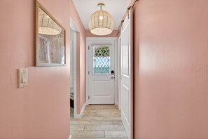 Un couloir aux murs roses, une porte blanche ornée d’un panneau de verre décoratif et un luminaire suspendu au plafond, fait de fibres tressées.
