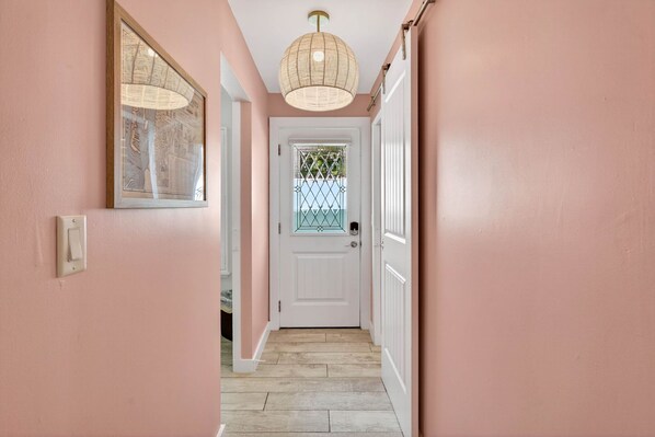 A hallway with pink walls, a white door with a decorative glass panel, and a woven ceiling light fixture.
