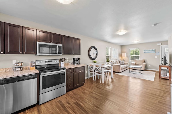 A modern kitchen with stainless steel appliances, a dining area with a white table and chairs, and a living room with a sofa and a TV. - Full Kitchen, In-Suite Laundry, Wi-Fi, and Parking (Wasilla)