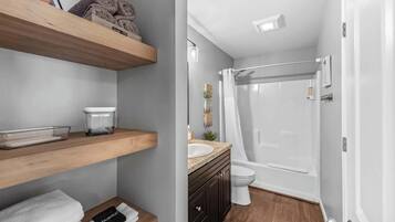 A bathroom with a shower, toilet, and vanity. There are wooden shelves with towels and a glass container.