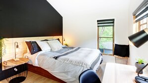 Ett modernt sovrum med ett sluttande tak, en säng med ett svart- och vitrandigt överkast, ett sängbord med en lampa och ett fönster med utsikt över grönområden.