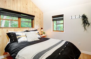 A bedroom with a large bed, wooden walls, two windows, a bedside table, a lamp, and a hanging plant.