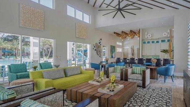 Amplia sala de estar con un sofá verde brillante, sillas azules y una mesa de centro de madera. Se observa un área de cocina con sillas azules y una mesa de comedor. La habitación tiene ventanas grandes y un diseño de techo moderno.