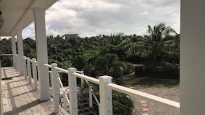 A wooden deck with white railings overlooking a lush garden with palm trees and a building in the background. - Private Home on Amazing Beach! 22 Steps to Beach!  (Coconut Bay)