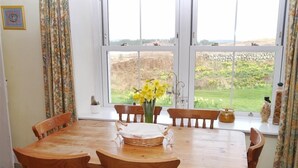 A dining room with a wooden table and chairs, a vase of yellow flowers, and a view of a garden through large windows. - Airie Farmhouse -  a house that sleeps 6 guests  in 3 bedrooms (Castle Douglas)