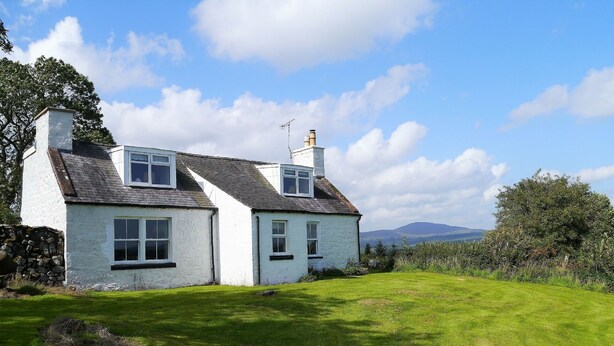 A two-story white house with a stone foundation, a grassy yard, and a mountain in the background. - Airie Farmhouse -  a house that sleeps 6 guests  in 3 bedrooms (Castle Douglas)
