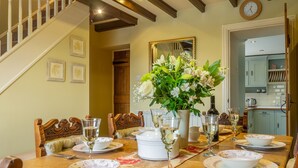 Uma mesa de jantar posta para seis pessoas, com louças brancas florais, um arranjo central branco e um relógio na parede.