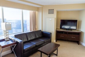 Ein Hotelzimmer mit einer Ledersofa, einem kleinen Couchtisch, einem Fernseher auf einem Holzständer und einem großen Fenster mit Blick auf die Stadt.