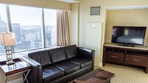 Habitación de hotel con sofá de cuero, mesa de centro de madera, televisor sobre un mueble de madera, escritorio con lámpara y un ventanal con vistas a la ciudad.