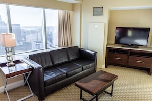 A hotel room with a leather sofa, a wooden coffee table, a TV on a wooden stand, a desk with a lamp, and a large window offering a city view.