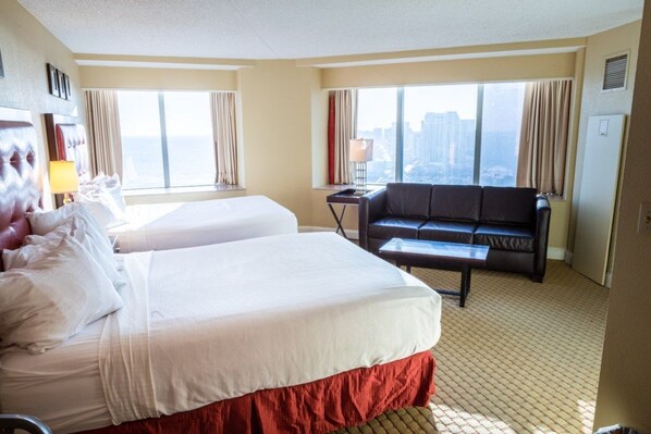 Une chambre d’hôtel avec deux lits, un canapé, un bureau et de grandes fenêtres donnant sur la ville.