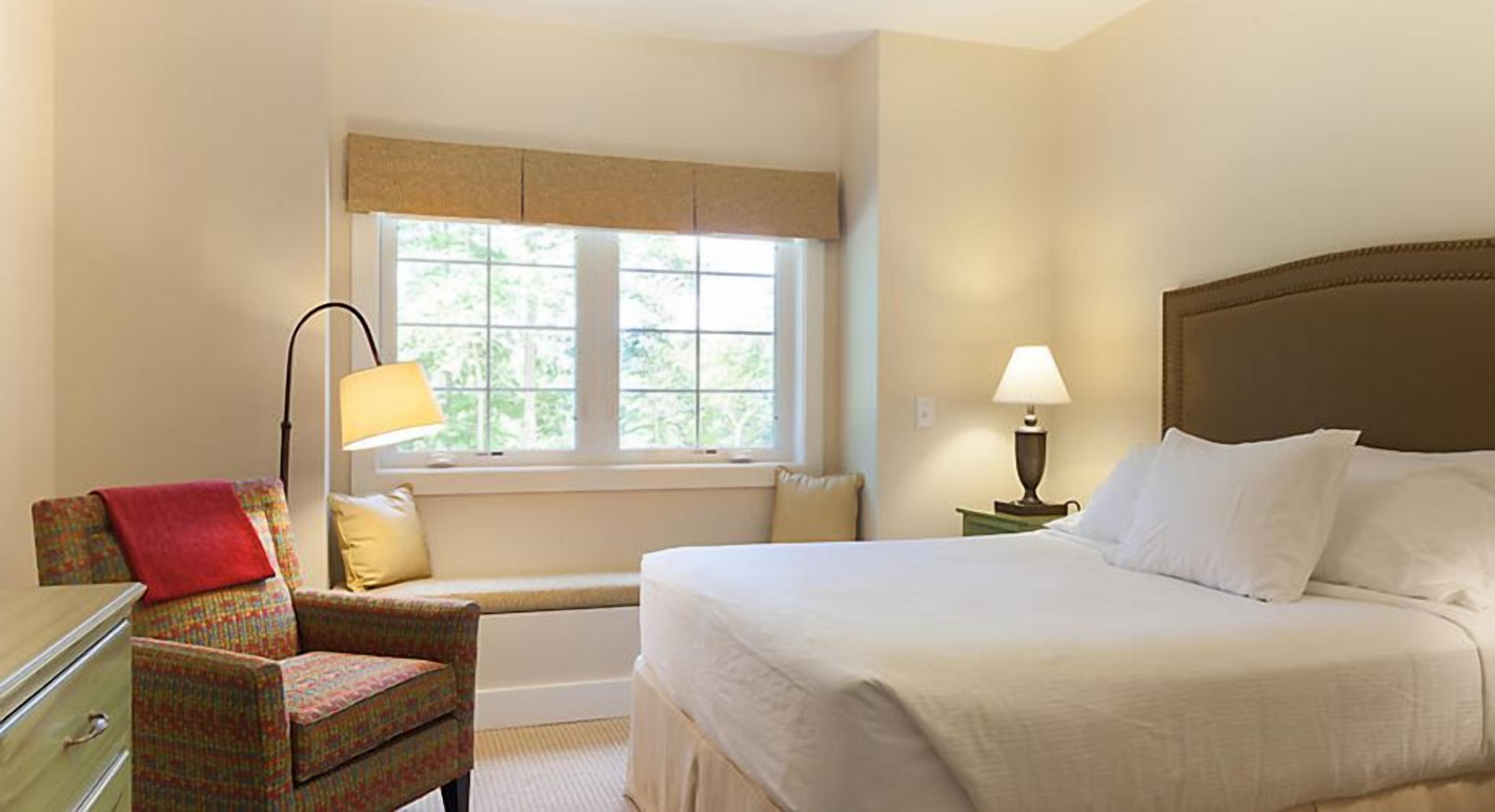 A hotel room with a bed, a chair, a window, and a lamp.