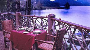 Un tavolo da pranzo apparecchiato con piatti, bicchieri e un vaso di fiori su un pontile in legno con vista su un lago.