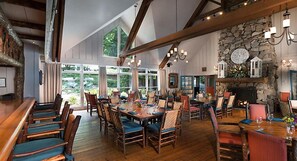 A spacious dining area with wooden tables and chairs, large windows, and a stone fireplace. - Cashiers | Family Nature Escape | Golf + Lake | 2BR (Cashiers)