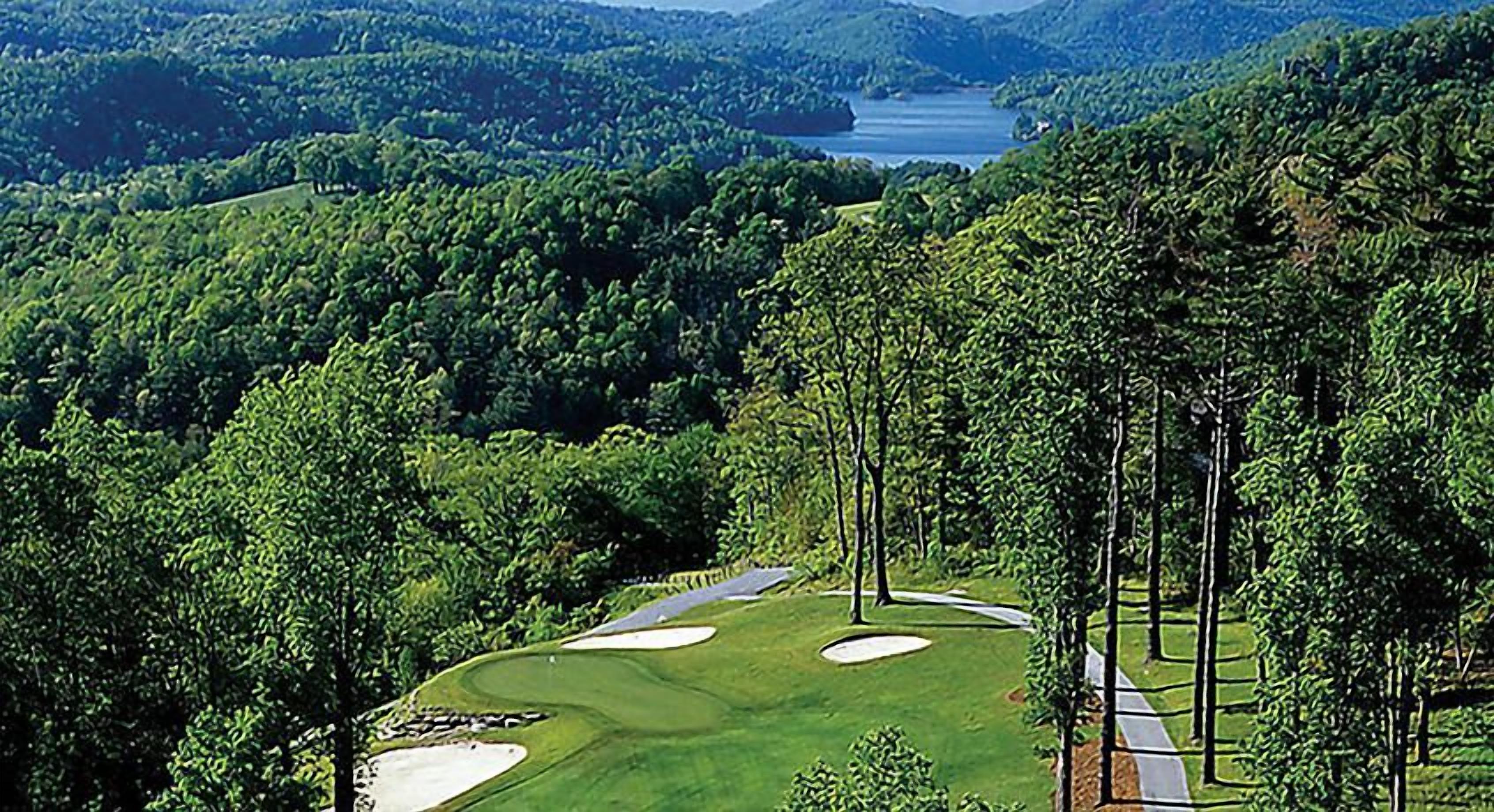 Ein Golfplatz mit Sandbunkern, einem Wasserkörper im Hintergrund und dichten, bewaldeten Hügeln.