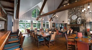 A spacious dining area with wooden tables and chairs, large windows, and a stone fireplace. - Cashiers | Premium Group Retreat | Golf + Lake | 3BR Sleeps 8 (Cashiers)