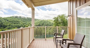 A wooden deck with a white railing, two wicker chairs, and a view of a green forested hill.