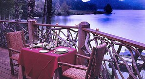 A dining table set with plates, glasses, and a vase of flowers on a deck with a wooden railing overlooking a lake. - Cashiers | Premium Group Retreat | Golf + Lake | 3BR Sleeps 8 (Cashiers)