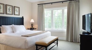 A hotel room with a large bed, a bedside table with a lamp, a bench, a TV, and a window with curtains. - Cashiers | Premium Group Retreat | Golf + Lake | 3BR Sleeps 8 (Cashiers)