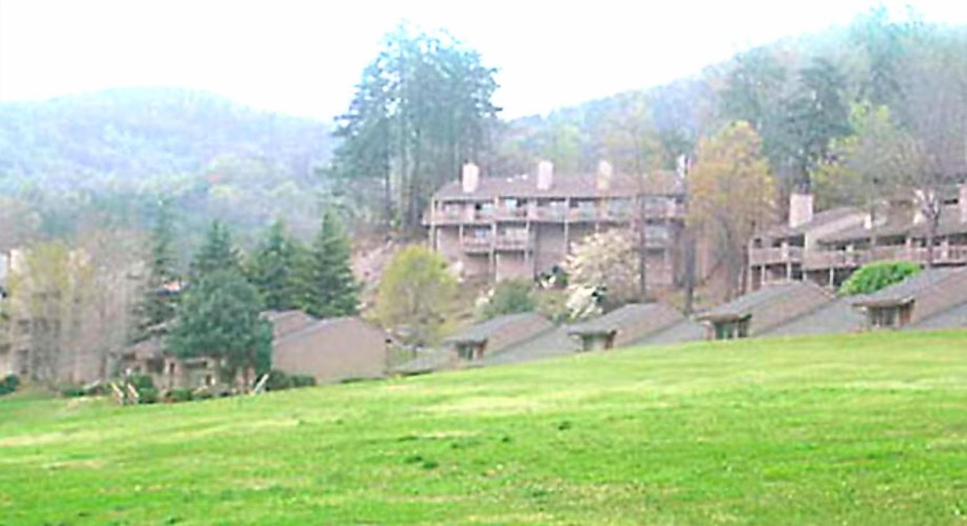 Um edifício com vários andares e telhado plano, cercado por árvores e um campo gramado.