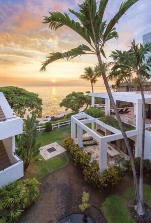 A coastal property with a view of the ocean at sunset, a white building, and palm trees. - Kona | Couples Hawaii Coast Stay | Pool + Views (Kailua-Kona)