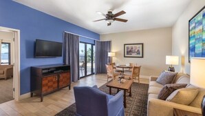 Ein Hotelzimmer mit Flachbildfernseher, Deckenventilator, Essbereich mit Tisch und Stühlen, einer Couch und einem großen Fenster mit Vorhängen.