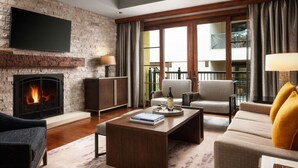A living room with a fireplace, a flat-screen TV mounted on a brick wall, a sofa, a coffee table with a book, and a view of a balcony with outdoor furniture.