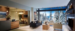 A modern living room with a sofa, coffee table, and a TV. There is a wooden reception desk and a dining area with chairs. The room has large windows with a view of snow-covered trees.