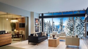 A modern living room with a sofa, coffee table, and a TV. There is a wooden reception desk and a dining area with chairs. The room has large windows with a view of snow-covered trees. - Ritz Carlton Club, Lake Tahoe - 4 Bedroom (Truckee)