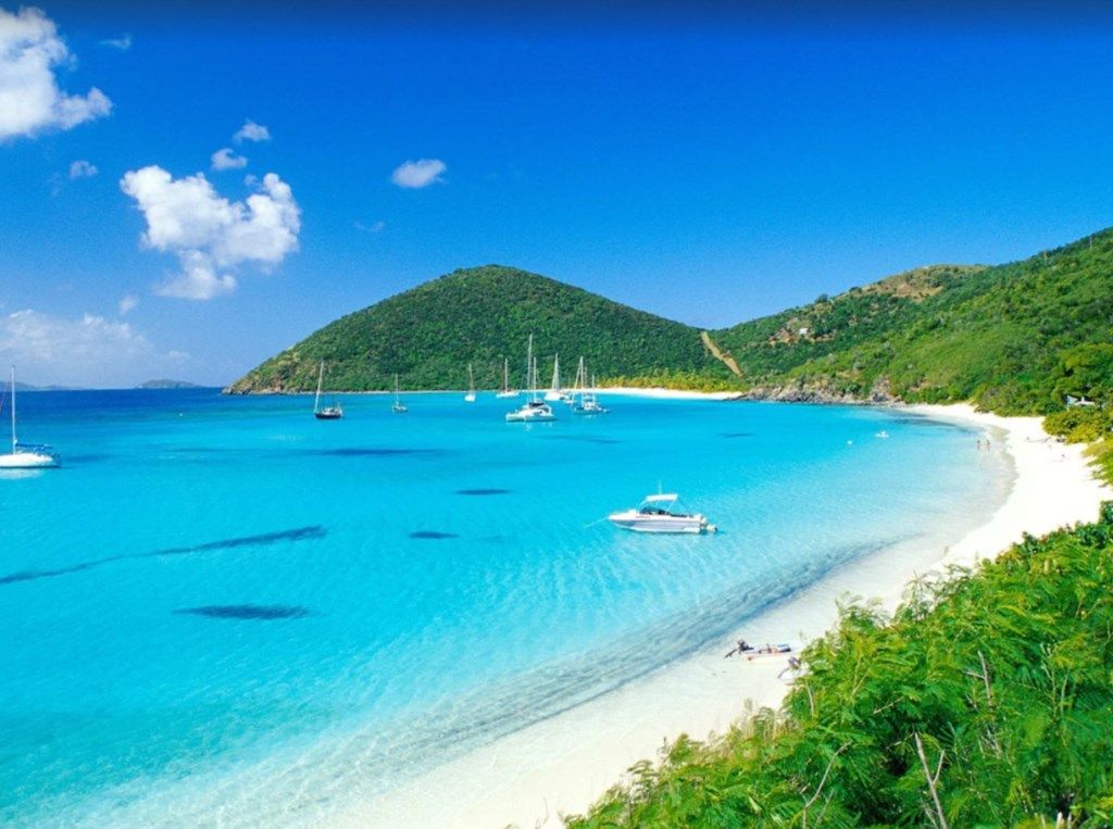 Une plage aux eaux turquoise et limpides, avec des bateaux et une chaîne de collines en arrière-plan.