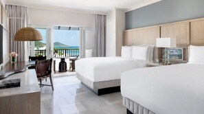 A hotel room with two beds, a desk, a chair, and a balcony with a view of the sea.