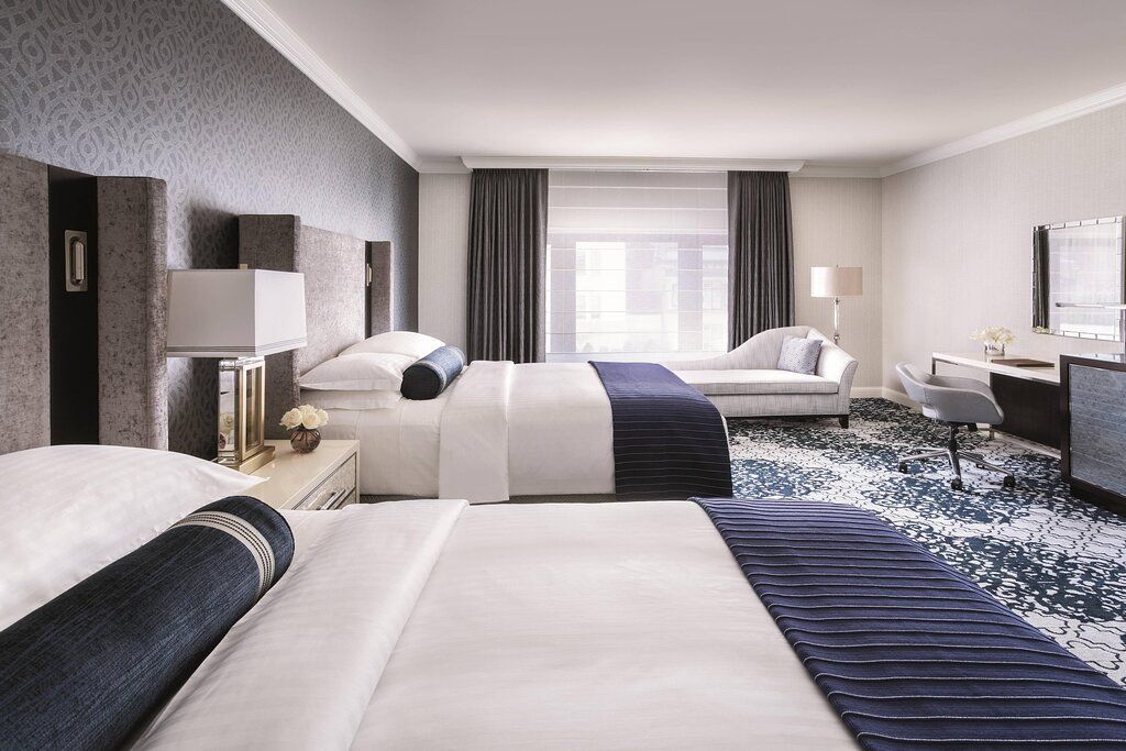 A hotel room with two beds, a desk, a chair, and a large window with curtains.