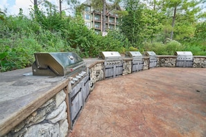 Un espace barbecue en pierre, doté de plusieurs grils, entouré de verdure et avec un bâtiment en arrière-plan.