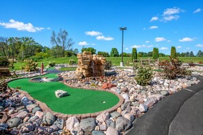 Un campo de mini golf con una superficie de césped verde, una formación rocosa como obstáculo y una valla de madera al fondo.