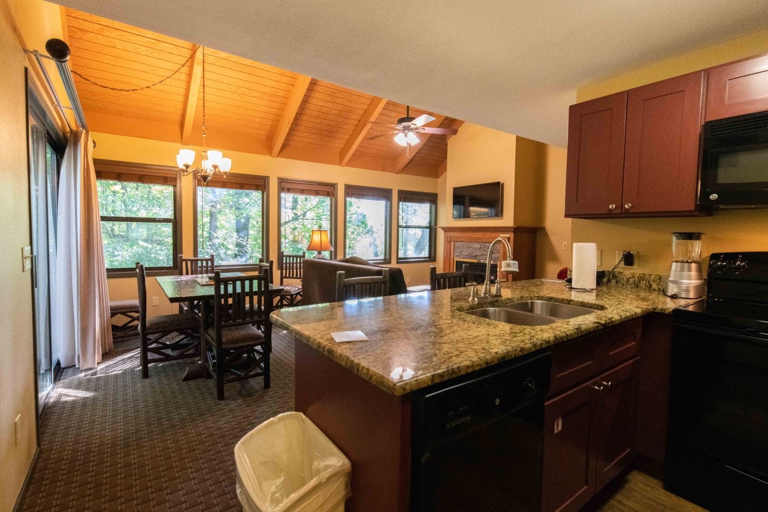 A kitchen with granite countertops, a dishwasher, a stove, and a microwave. There is a dining area with a table and chairs, and a window with a view of trees.