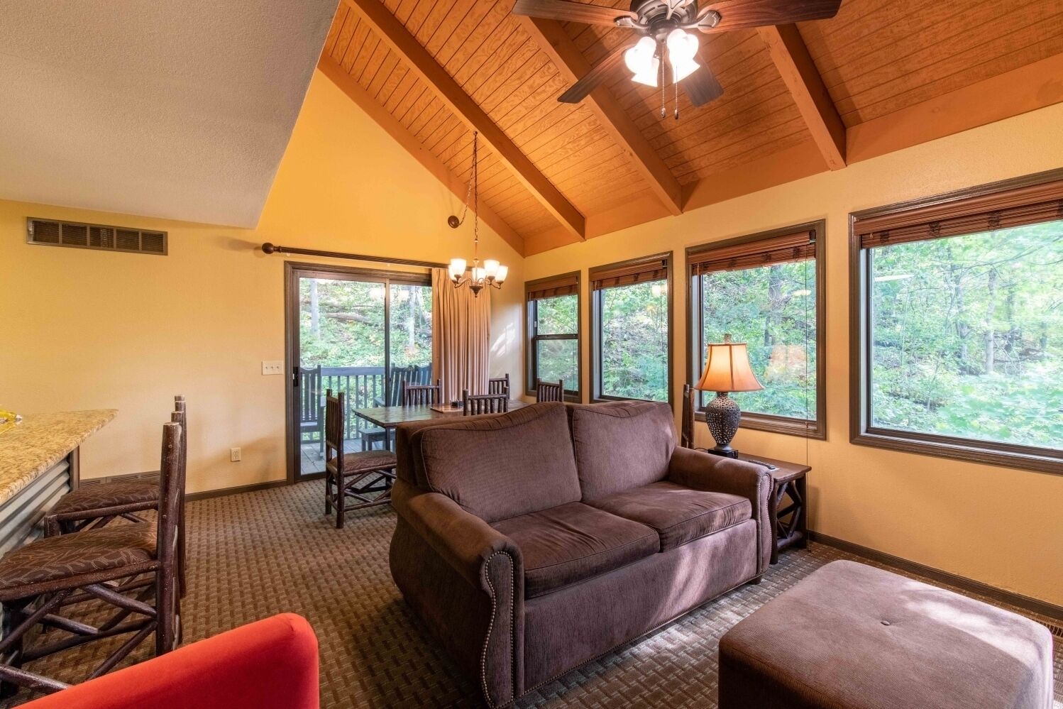 A cozy room with a brown leather sofa, a dining table with chairs, a wooden ceiling fan, and large windows offering a view of trees.
