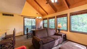 A cozy room with a brown leather sofa, a dining table with chairs, a wooden ceiling fan, and large windows offering a view of trees.