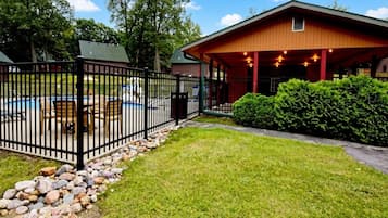 A fenced outdoor area with a pool, a covered seating area, and a building with a sloped roof.