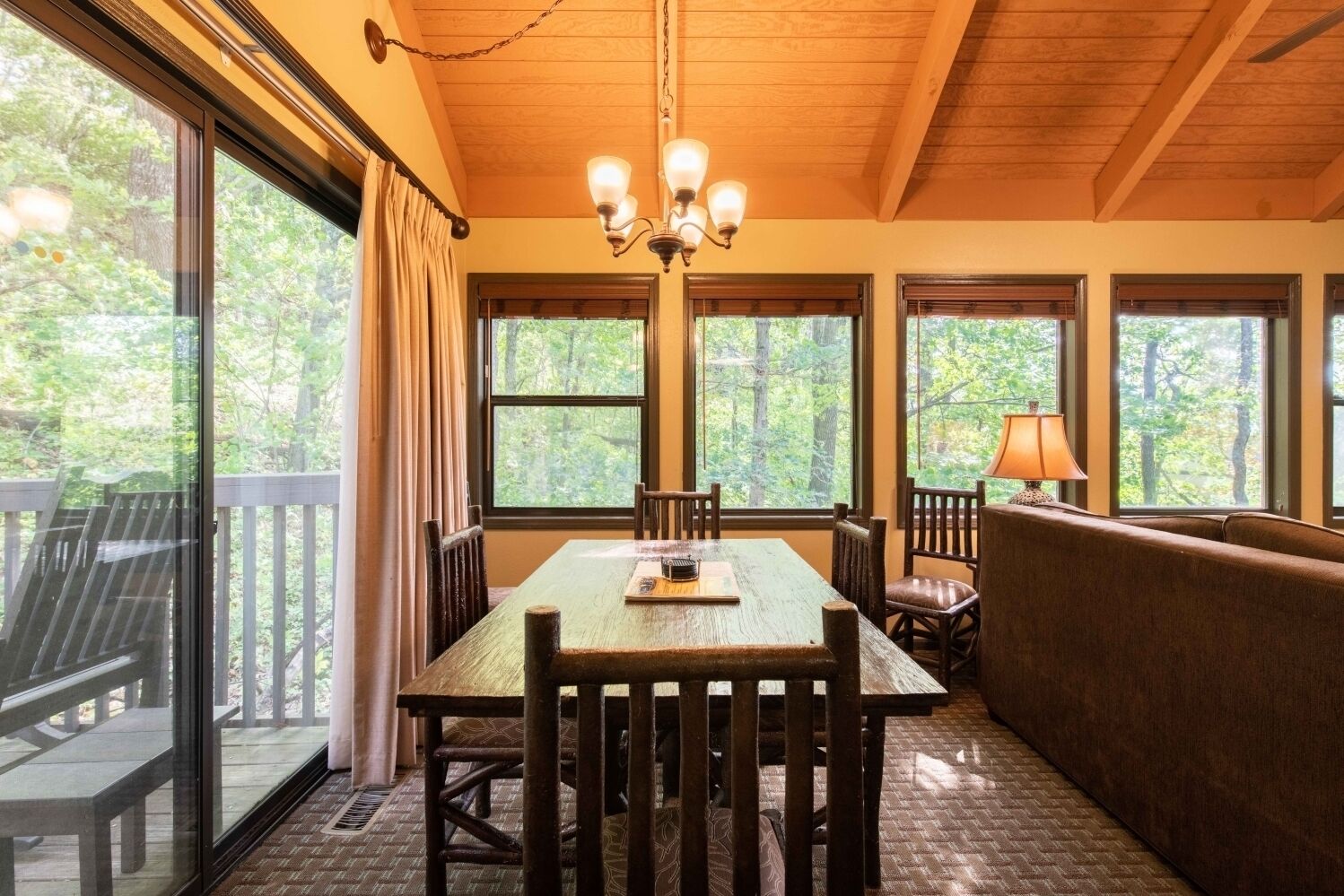 A dining area with a wooden table, chairs, and a bench. There are large windows with curtains, a chandelier, and a lamp on the table.