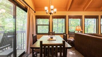 A dining area with a wooden table, chairs, and a bench. There are large windows with curtains, a chandelier, and a lamp on the table.