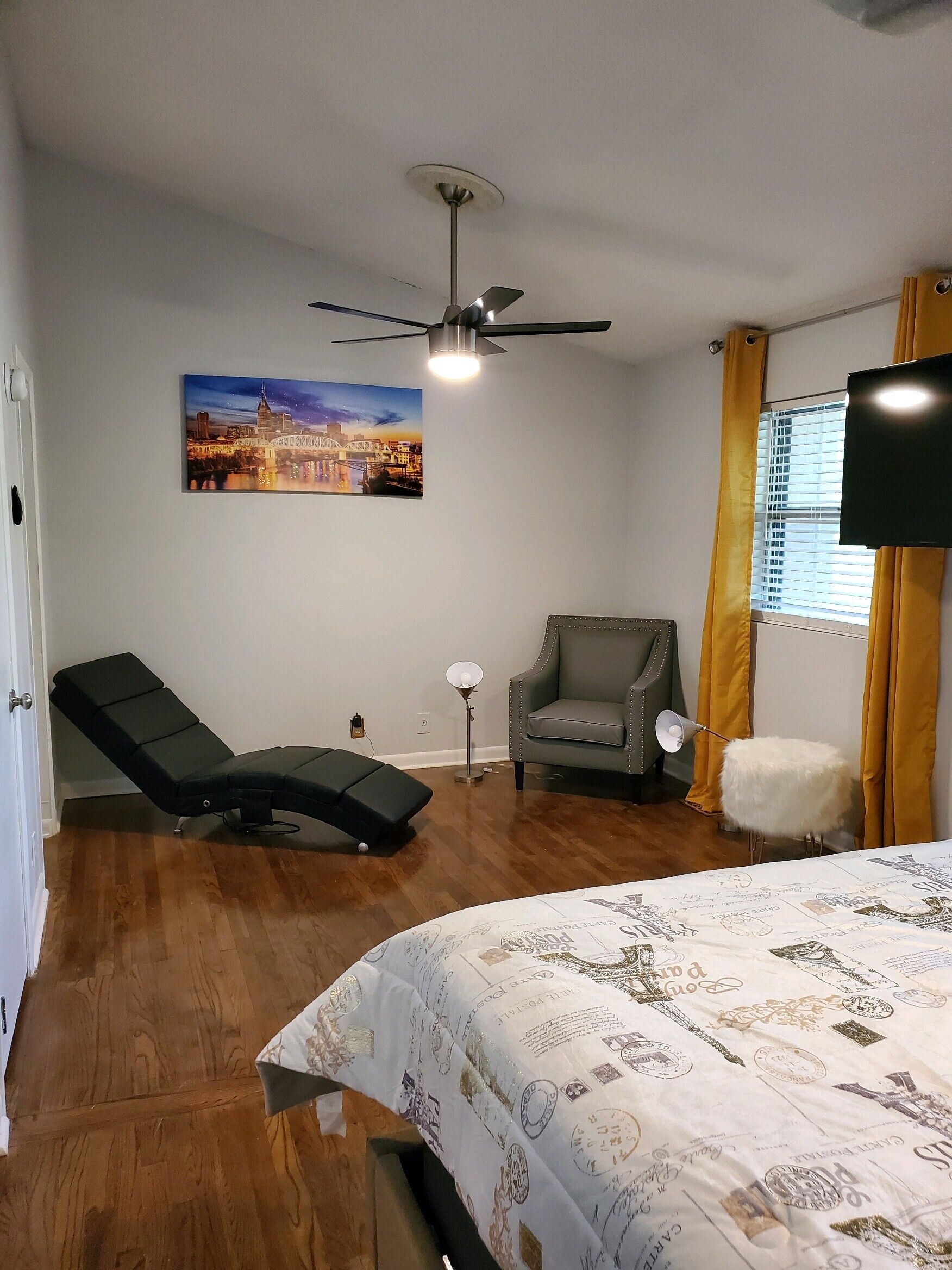 Ein Zimmer mit einem Bett, einer Massageliege, einem Stuhl, einem Deckenventilator und einem Fenster mit Vorhängen.