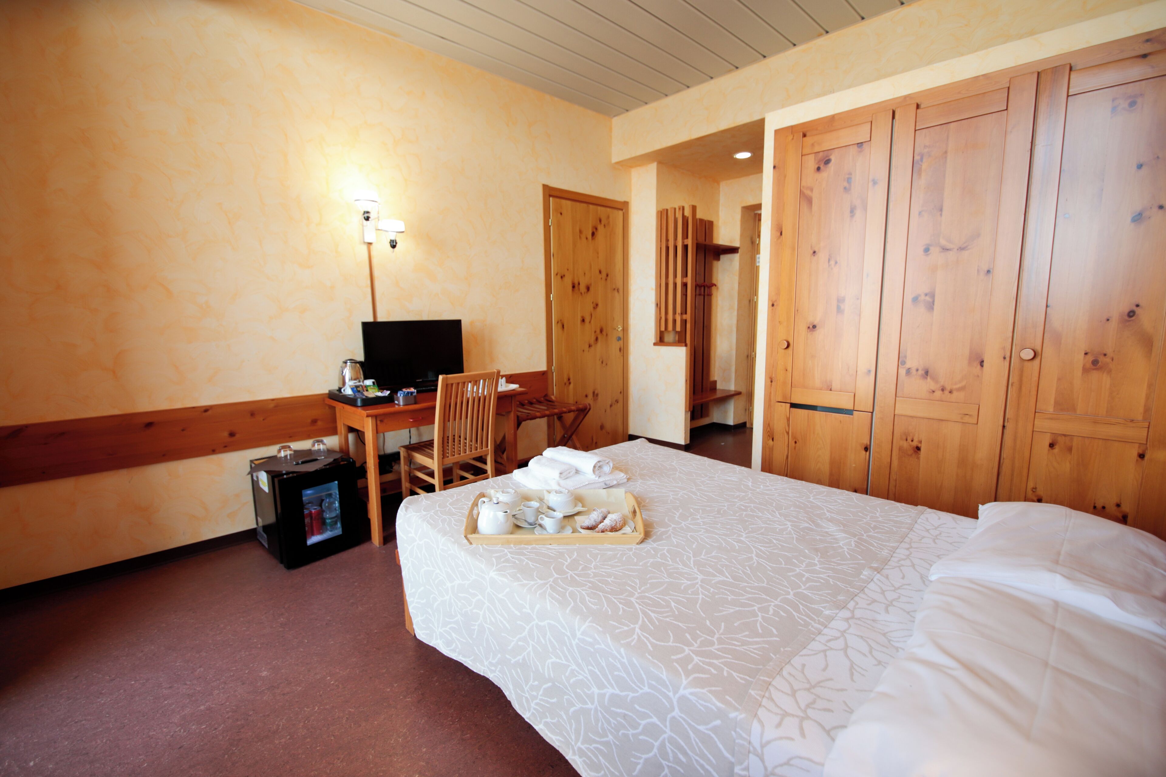 A hotel room with a bed, a desk with a computer, a chair, a tray with breakfast items, and wooden wardrobes.