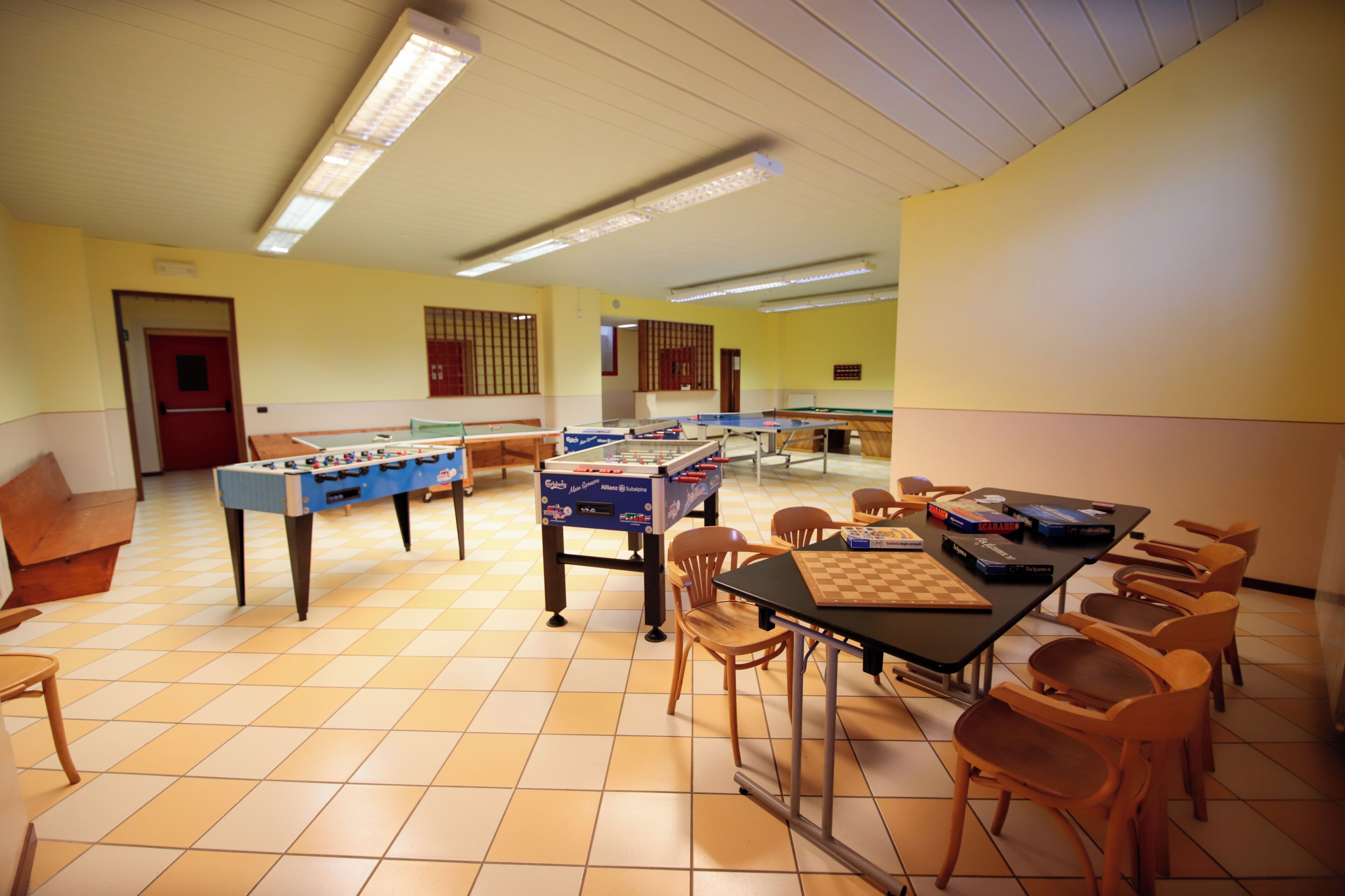 Una stanza con due tavoli da calcio balilla, un tavolo da ping pong e un tavolo da scacchi. Ci sono diverse sedie e un tavolo con dei libri.